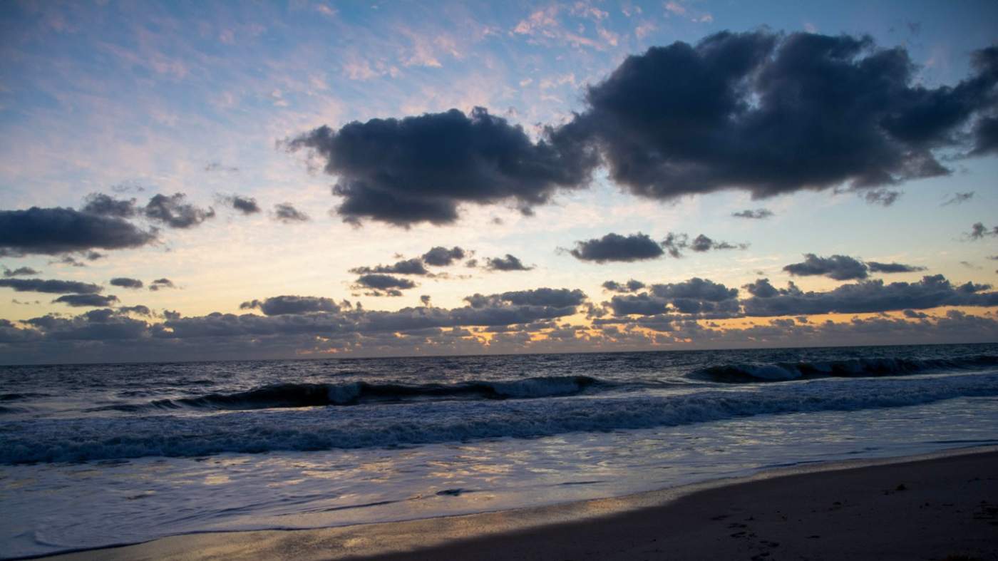 A tranquil beach at sunrise. The sun casts a warm glow across the sky, reflecting off the water. Gentle waves lap onto the sandy shore. Partly cloudy sky with dark, dense clouds contrasting with lighter shades of blue and orange.