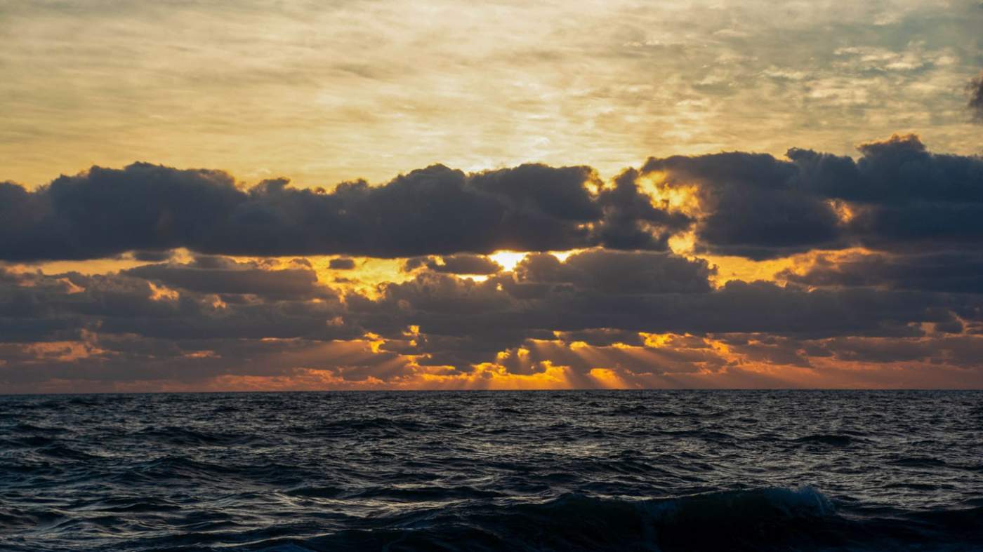 A tranquil seascape at sunrise with the sun partially obscured by cumulus clouds. The sunlight diffuses, creating a warm golden glow that reflects off the choppy ocean waves. The horizon is visible, separating the dark blue water from the orange and yellow hues of the sky, suggesting a peaceful start to the day.