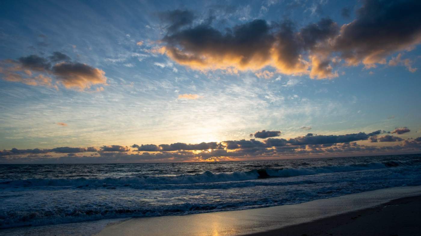 A scenic view of a beach at sunrise with the sun partially visible on the horizon. The sky is filled with scattered clouds, some of which are underlit by the sun’s glow, creating a dramatic contrast. The ocean waves are gentle and rolling onto the sandy shore, reflecting the sunlight in patches. The image captures the tranquil beauty of a coastal landscape under changing light conditions.