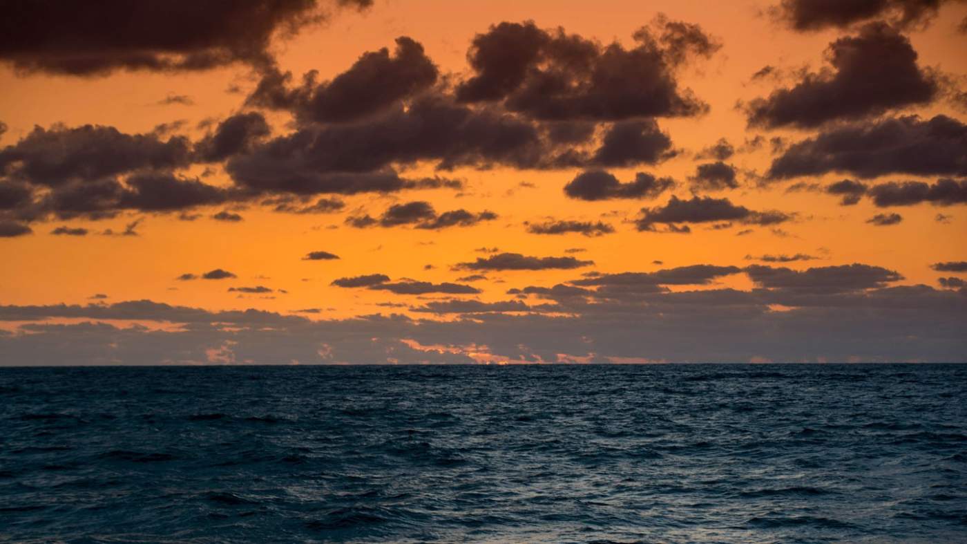 A serene seascape at sunrise: The vibrant display of orange and yellow hues in the sky is dotted with numerous dark clouds. The calm sea reflects the colors of the sky, creating a harmonious blend of warm tones over the water’s surface