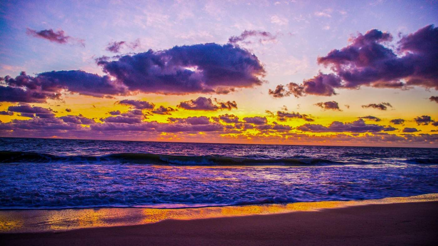 A vibrant beach scene at sunrise with a dynamic sky filled with purple and orange hues reflecting on the ocean waves, while the sun rises near the horizon line. The beach in the foreground shows wet sand, indicating the tide’s movement.