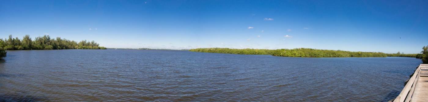 A calm, wide river with rippling water flows under a clear blue sky. Green trees line the far bank in the distance.