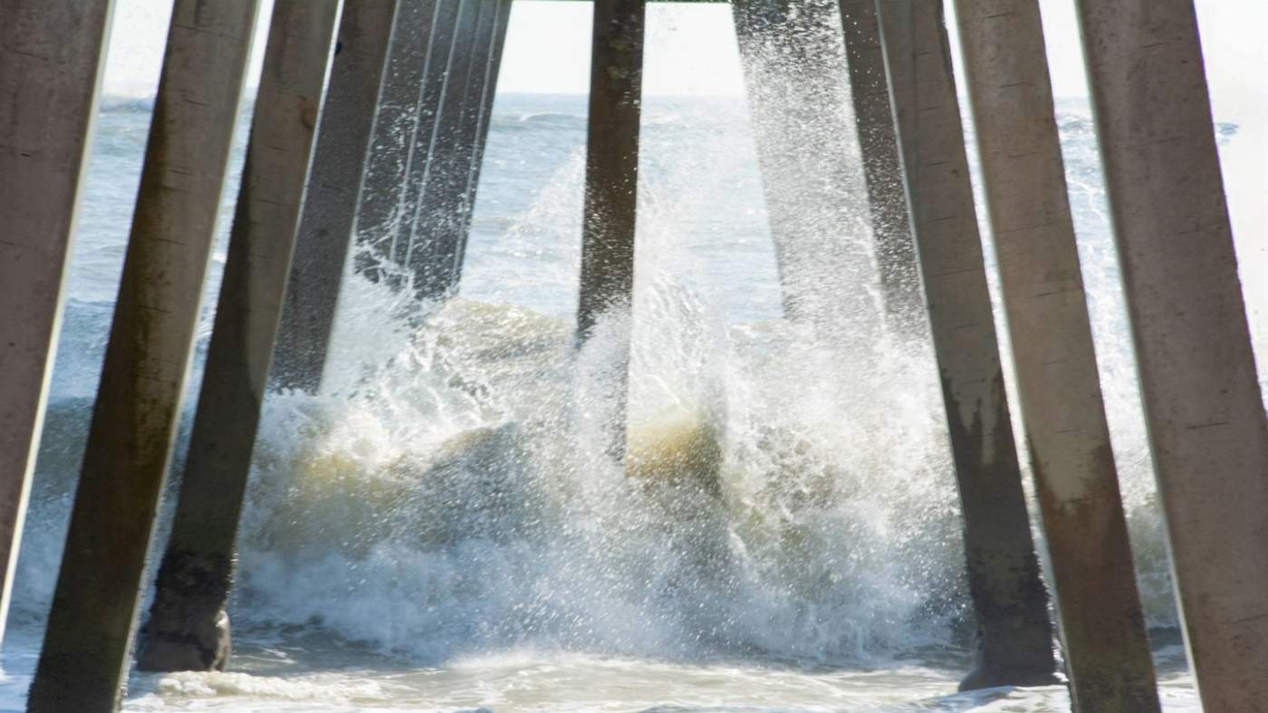Waves crashing against the pillars of a pier, causing splashes of water. Sunlight filters through the gaps, creating a dynamic interplay of light and shadow in the scene.