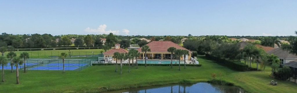Aerial view of the red-tiled roof, of the backside of the Citrus Springs Clubhouse, surrounded by a manicured lawn and palm trees. To the left, there are tennis courts. In the foreground, a calm pond reflects the blue sky and clouds.