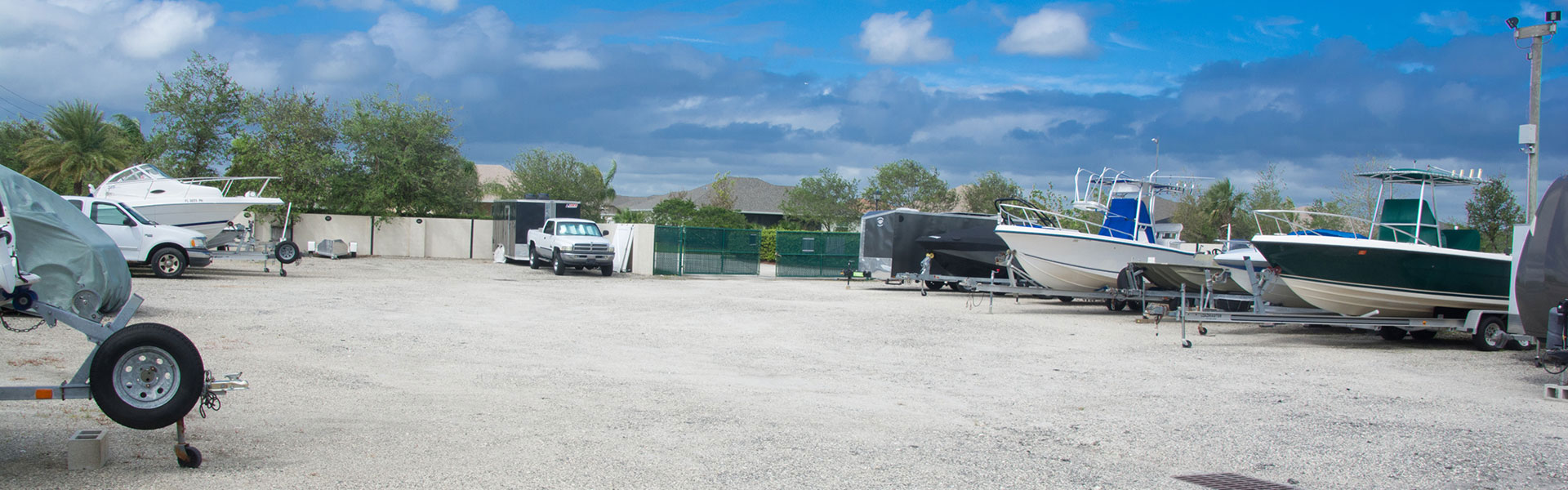 Several boats and trailers are parked on a gravel lot under a partly cloudy sky. Trees and a truck are visible in the background.
