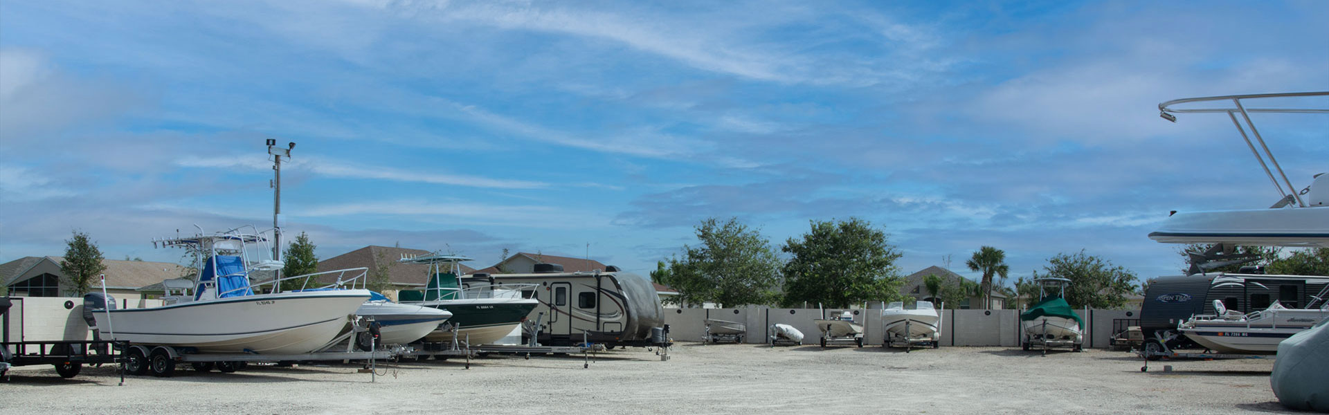 The Citrus Springs RV lot with various boats, trailers, and RVs parked on a gravel surface under a blue sky with clouds. Fenced area in the background with trees and houses visible.