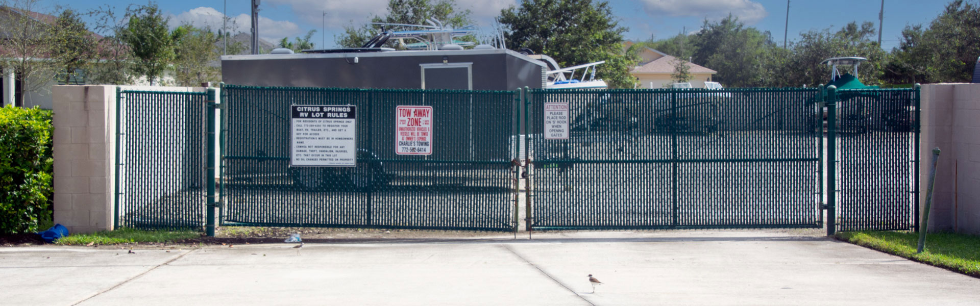 The gated entrance to the Citrus Springs RV lot with signs displays rules and contact information. Behind the fence, a trailer is seen, parked on a concrete lot. Trees and a clear blue sky are visible in the background.