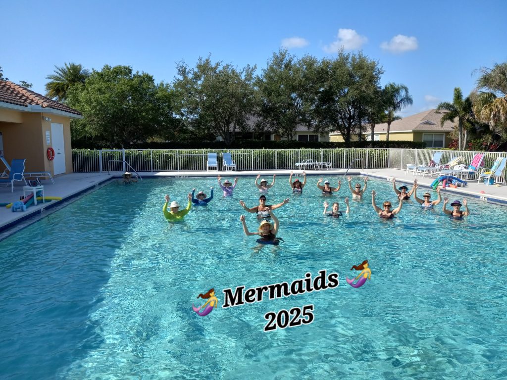 A group of people in a pool pose with raised arms, forming a circle. Text on the water reads "Mermaids 2025" with mermaid emojis. Lounge chairs and trees are in the background under a blue sky.