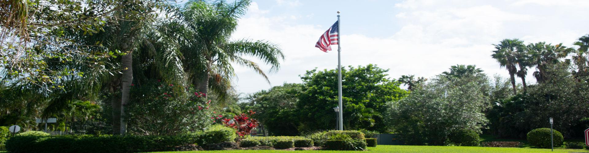 A sunny park scene featuring an American flag waving on a flagpole, surrounded by lush greenery, including palm trees and shrubs. The sky is partly cloudy, creating a serene and peaceful atmosphere.