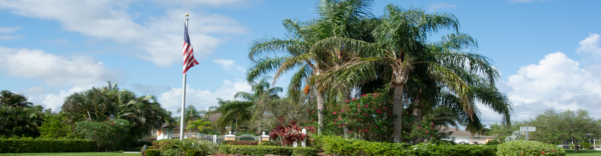 A scenic view of a landscaped area featuring a tall flagpole with an American flag. Lush palm trees and bushes with red flowers are in the foreground, while houses and a clear blue sky with scattered clouds are in the background.