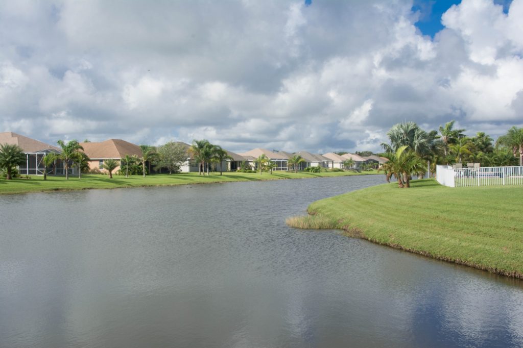 A serene suburban lake with a grassy shoreline, lined with palm trees and houses under a partly cloudy sky.